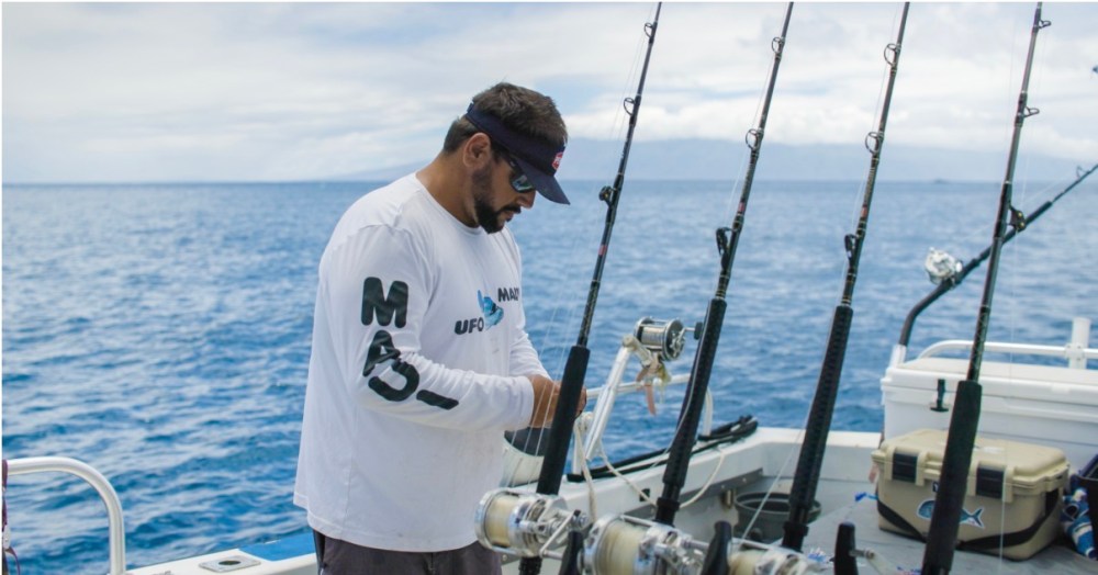 Man on boat with fishing rods, ocean in background, wearing a visor and long-sleeve shirt.